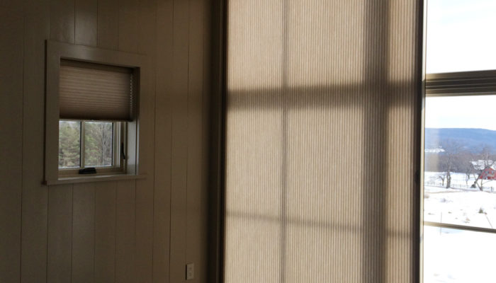 This Is A Picture Of A Wall With A Small Window Which Features Light Filtering Shades And A Large Window Perpendicular To It Which Features Vertical Blinds.