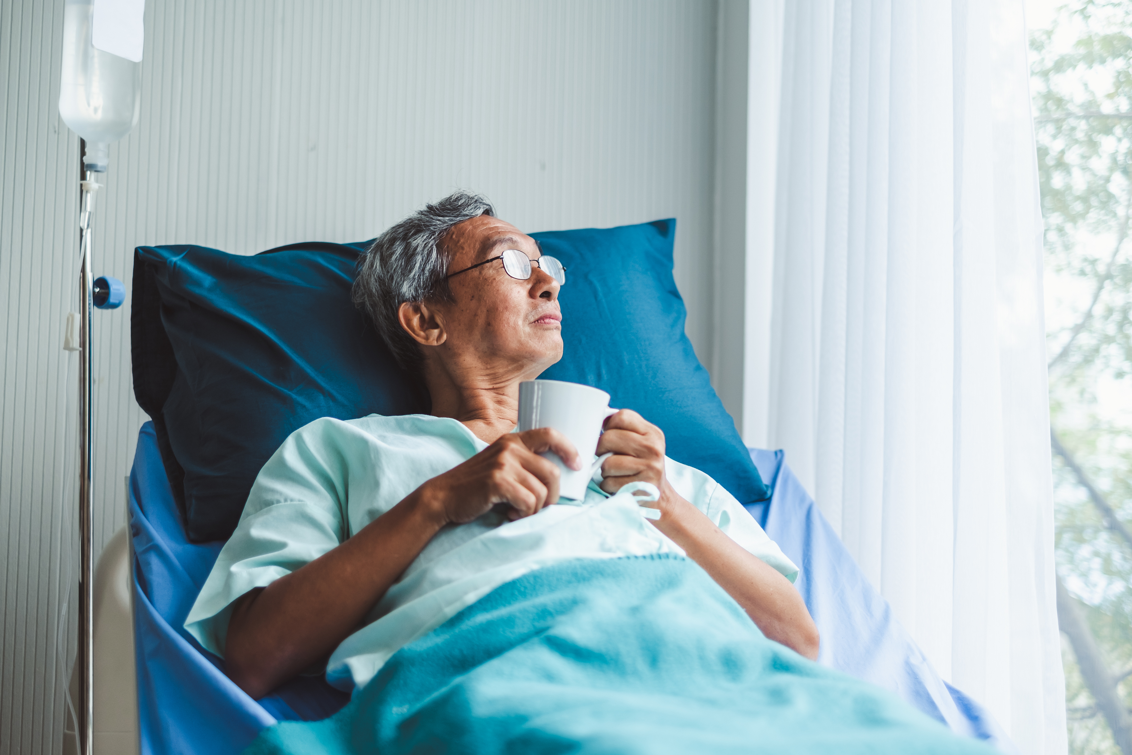 This is a picture of an elderly Asian man lying in a hospital bed looking out the window which features vertical blinds.